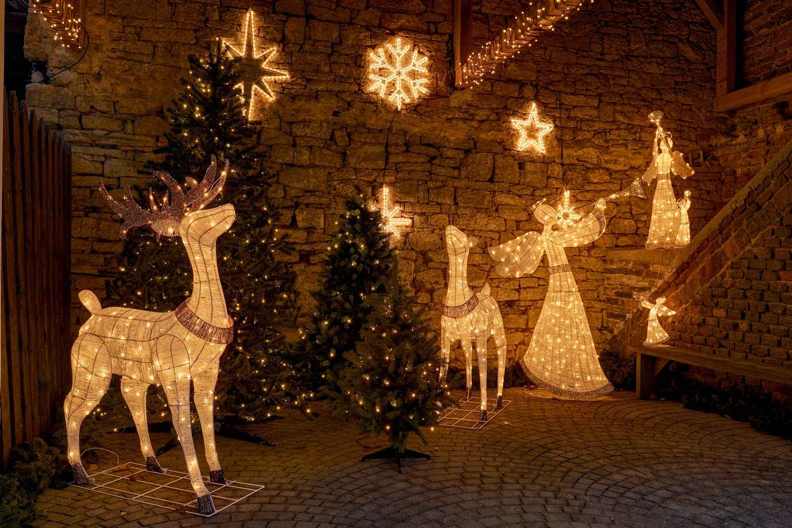 Udstilling af julebelysning med lysende rensdyr, engle og stjerner mod en rustik stenvæg.